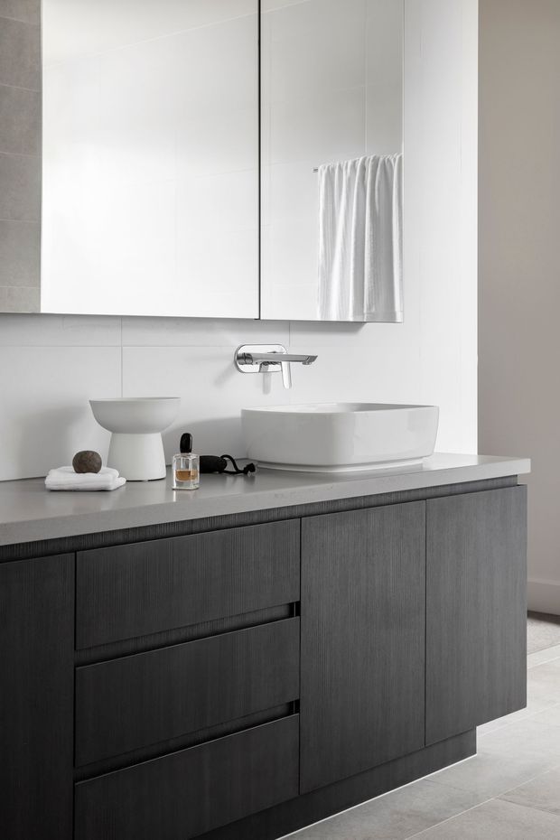 A bright, contemporary bathroom vanity featuring a wall-mounted mirror cabinet, white vessel basin, chrome wall-mounted tapware, light grey benchtop, and dark vertically grained timber cabinetry. Large white wall tiles and pale floor tiles create a clean, minimalist fit-out.