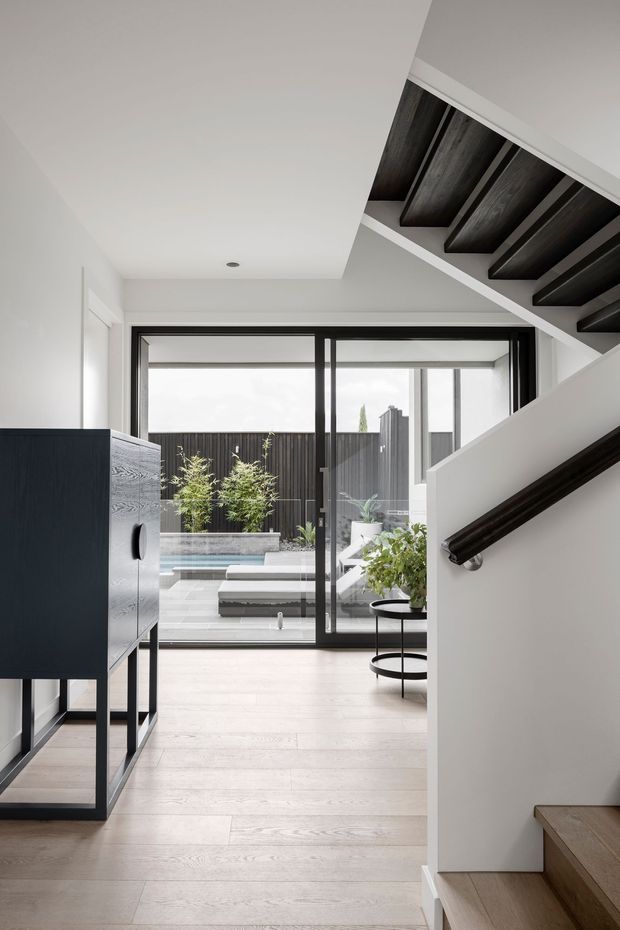 A bright, refined residential hallway and stairway viewed from inside. Pale timber flooring and white walls lead toward full-height black-framed glazing opening onto a compact courtyard with a pool. A dark timber console stands on the left, while the staircase on the right features pale timber treads, a dark handrail, and exposed dark timber underside beams.