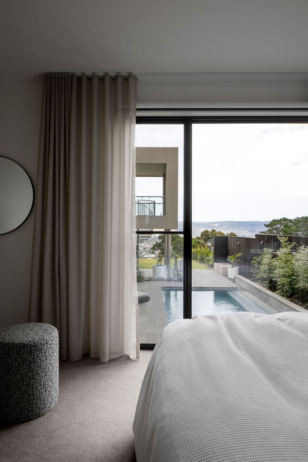 A contemporary bedroom with a soft neutral palette, floor-to-ceiling curtains, grey carpet, and a partially visible bed dressed in white textured bedding. Full-height black-framed glazing opens to a terrace and narrow swimming pool, with landscaped planting and a distant coastal or suburban view beyond. A round wall mirror and textured cylindrical stool add understated furnishings.