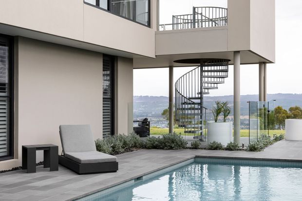 Contemporary multi-level home exterior with a cantilevered upper floor, slender structural columns, glass balustrades and a prominent external spiral staircase. A swimming pool and paved terrace occupy the foreground, with landscaped planting and a distant elevated landscape visible beyond.