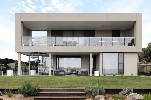 Contemporary two-storey residence with a strong rectilinear form, deep upper-level balcony, glass balustrade, and extensive glazing. The lower level opens to a landscaped lawn and patio, while a prominent white spiral staircase connects to the balcony. Neutral rendered finishes, dark window frames, and clean minimalist lines define the facade.