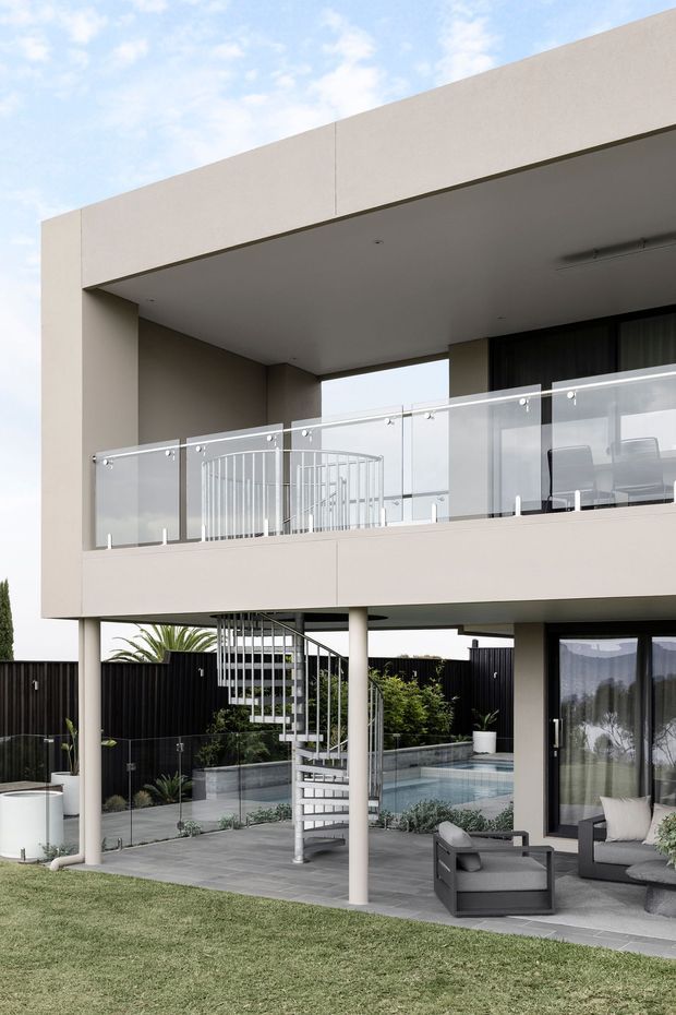 Exterior view of a contemporary two-storey home with a projecting upper-level terrace, smooth light render, glass balustrades, dark-framed glazing, and an open ground-level outdoor living area. A spiral staircase connects the terrace and paved patio beside a swimming pool, with minimalist outdoor furniture and landscaped lawn.
