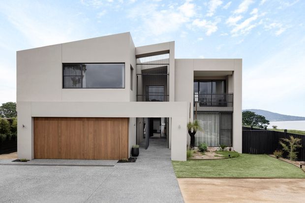 Street-facing view of a contemporary two-storey residence with a minimalist rendered facade, broad timber garage door, recessed entry, vertical screening, balconies, and large glazed openings. A landscaped front lawn and driveway frame the house, with a visible coastal or waterfront outlook in the background.