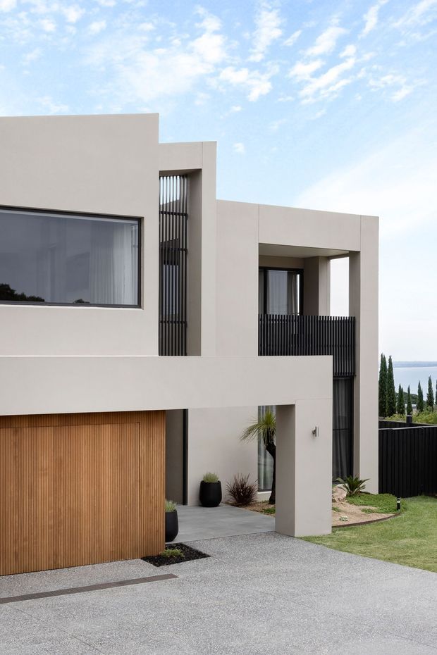 Contemporary multi-level residential facade with clean geometric volumes, smooth light render, dark vertical metal screens and balustrades, expansive glazing, and a warm timber garage door. A broad concrete driveway and minimalist landscaped entry lead toward the house, with distant water visible beyond the property.