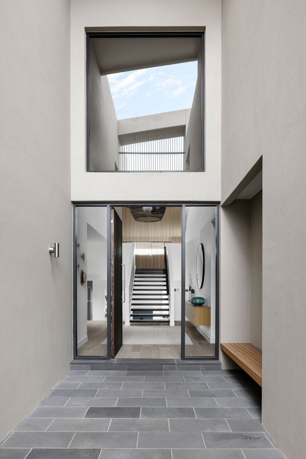 A refined modern entry courtyard viewed from outside, with a grey tiled approach leading to large glazed entry doors. The tall entry volume features a double-height opening, skylit void, slim dark window frames, white rendered walls, an internal staircase with bold horizontal treads, vertical timber screening, and a built-in timber bench along the right wall.
