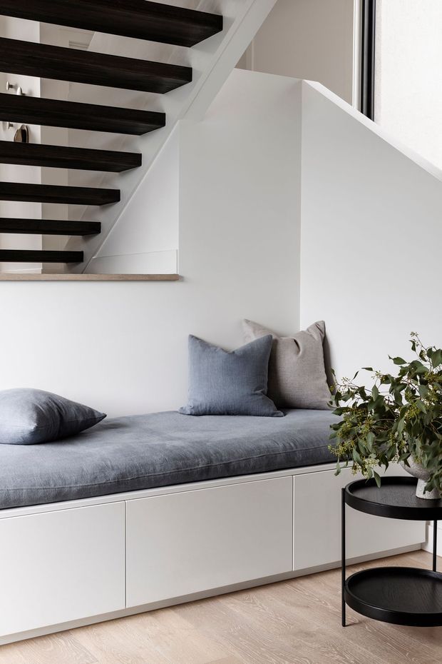 A bright, minimalist interior featuring a built-in window seat with pale cabinetry and a soft grey cushion, styled with grey and taupe pillows. Above it is an open staircase with dark timber treads and white-painted structural elements. A black round side table with a potted plant sits on a light timber floor beside the bench.