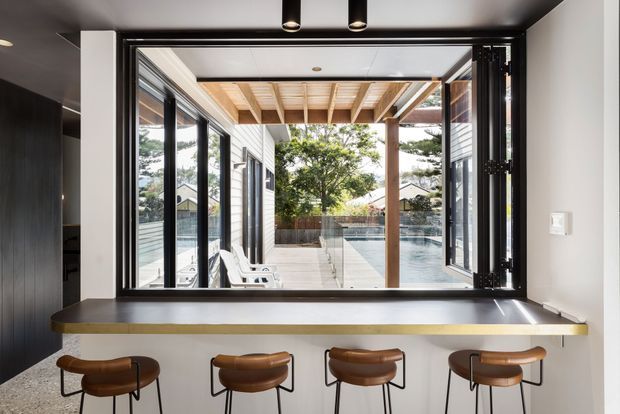 A contemporary kitchen or entertaining area with a long dark benchtop and four brown leather stools positioned at a large servery window. Fully opened black-framed glazing connects the interior to a covered timber outdoor deck and a swimming pool beyond. The interior has dark cabinetry, white walls, a dark ceiling, recessed spotlights, and strong indoor-outdoor flow.