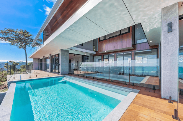 A contemporary multi-level outdoor entertaining area with a turquoise swimming pool in the foreground, timber decking, frameless glass balustrades, and expansive glazing beneath broad cantilevered roof planes. The building combines pale rendered or concrete columns, warm timber soffits and wall cladding, and dark window frames, with a bright coastal or hillside outlook beyond.