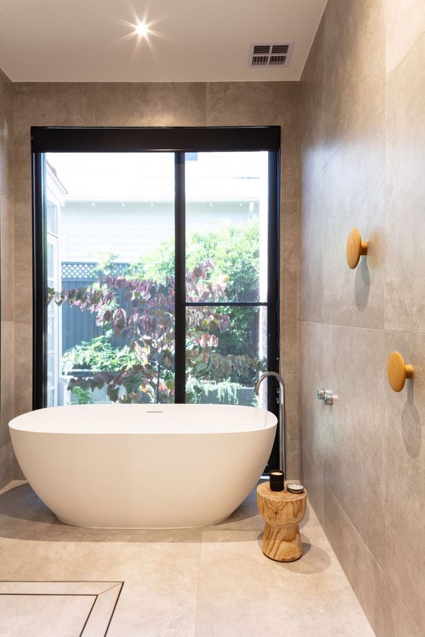A contemporary bathroom featuring a freestanding white soaking tub positioned in front of a large black-framed window. Large-format taupe stone-look tiles cover the walls and floor, with warm gold wall-mounted controls, a linear floor drain, recessed ceiling light and ventilation grille. A small sculptural timber stool sits beside the bath, with garden greenery visible outside.