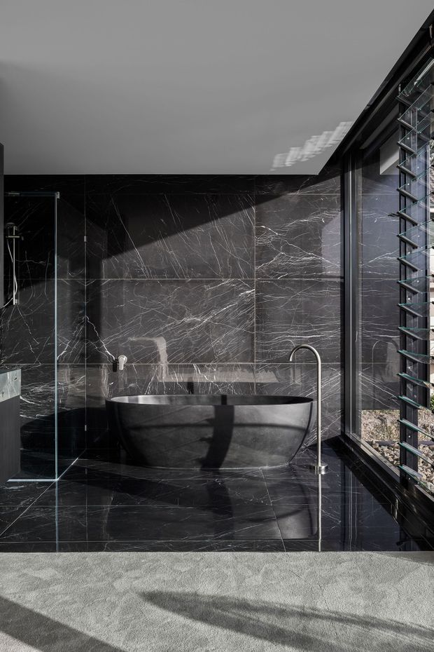 A dramatic contemporary bathroom featuring a freestanding matte-black bathtub, black marble-effect wall and floor surfaces with prominent white veining, a frameless glass shower enclosure, and a large louvred window providing strong natural light. The space has a dark, luxurious, high-contrast appearance.