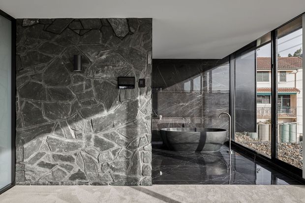 A luxurious contemporary bathroom featuring a freestanding dark stone bathtub beside floor-to-ceiling black-framed glazing. The room combines polished dark marble-like wall and floor surfaces with a rugged irregular stone feature wall, black fixtures, and a minimalist monochromatic palette. A neighbouring building is visible through the windows.