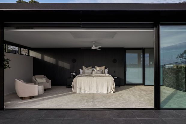 A contemporary bedroom opening directly onto an outdoor terrace through wide black-framed sliding glass doors. The room features a low-profile bed with layered neutral bedding, upholstered armchairs, dark vertical wall panelling, pale carpet, a white ceiling fan, and a recessed ceiling detail. The open glazing creates a strong indoor-outdoor connection, with a dark tiled terrace visible in the foreground and landscape glimpsed beyond.