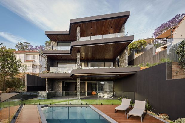 A multi-level contemporary residential house on a sloped suburban site, viewed from the rear garden. The home features dramatic projecting flat roof slabs, extensive glazing, dark cladding, timber-lined soffits and balconies with glass balustrades. A swimming pool with timber decking occupies the foreground, while mature landscaping and neighbouring homes surround the property.