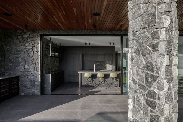 A dark, sophisticated covered outdoor entertaining area opens directly into a modern kitchen through large glazed sliding doors. The space features rough grey stone walls and a substantial stone-clad column, charcoal tiled flooring, a warm timber-lined ceiling with recessed lighting, and a compact kitchen island with stools visible inside.