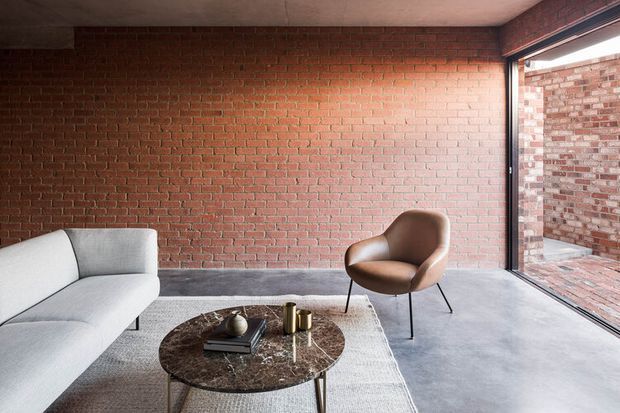 A minimalist living room with exposed red brick walls, a smooth concrete ceiling and floor, and full-height corner glazing opening to a brick courtyard. The room contains a light grey sectional sofa, a brown sculptural lounge chair, and a round dark stone coffee table over a neutral rug. The restrained material palette and industrial detailing create a refined, contemplative atmosphere.