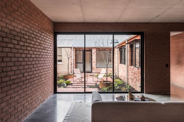 A refined brick-lined living room opens through expansive black-framed glazing onto a landscaped internal courtyard. The room has a polished concrete floor, exposed brick walls, a smooth concrete ceiling, and a neutral sectional sofa, creating a restrained, industrial-contemporary atmosphere. The courtyard beyond includes brick paving, low planting, outdoor chairs, and views of adjoining brick walls and corrugated roofing.