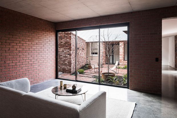 A modern living room with exposed red brick walls, a pale upholstered sofa, round glass coffee table, polished concrete flooring, and a large black-framed glazed opening overlooking a landscaped brick courtyard. A hallway or adjoining interior space is visible to the right.