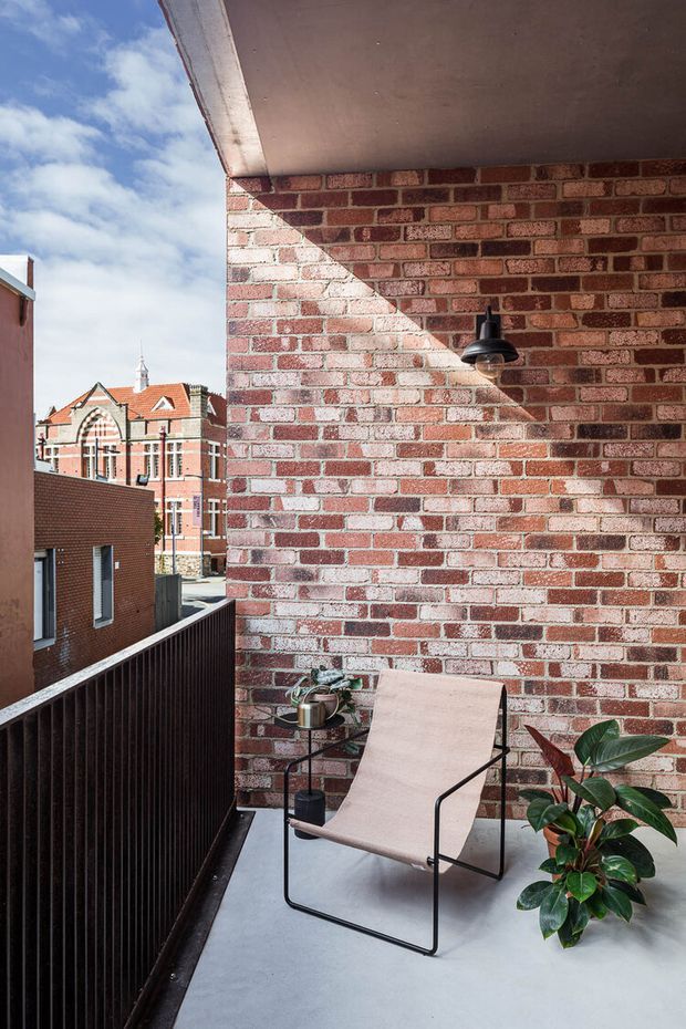 A compact urban balcony or terrace with a pale concrete floor, exposed red brick wall, dark vertical-slatted balustrade, and a simple sling lounge chair. Potted greenery and a small side table add warmth, while adjacent brick buildings and a historic gabled structure are visible beyond the balcony. A black wall-mounted light is fixed to the brick wall beneath a concrete overhang. The scene has a contemporary urban character with strong sunlight and clear blue sky.