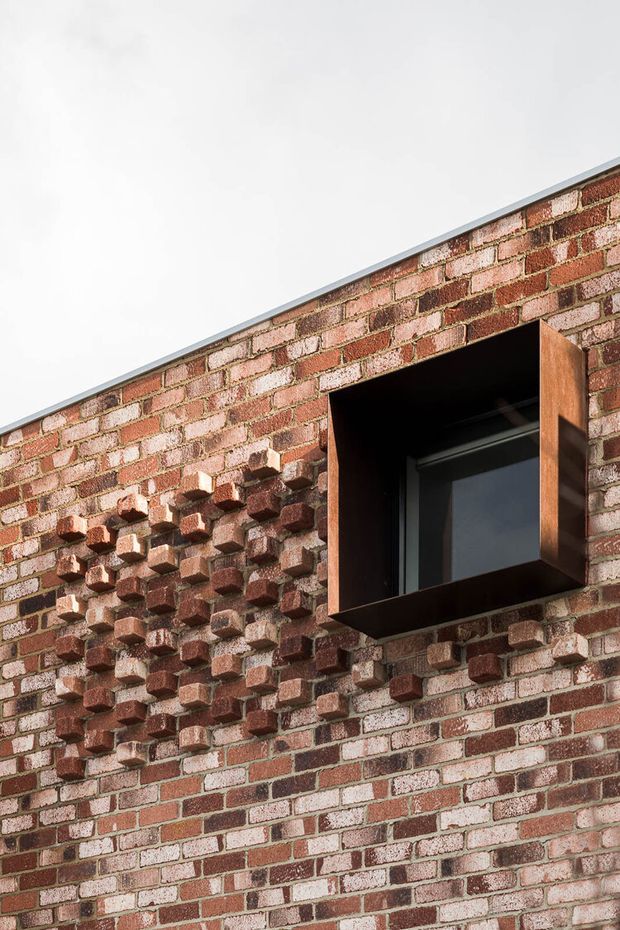 Close-up exterior view of a contemporary brick facade with deliberately projecting brickwork creating a textured geometric pattern. A dark-framed window is recessed within a deep, box-like timber-toned surround, and the wall has a minimal metal roof or parapet edge against a bright sky.