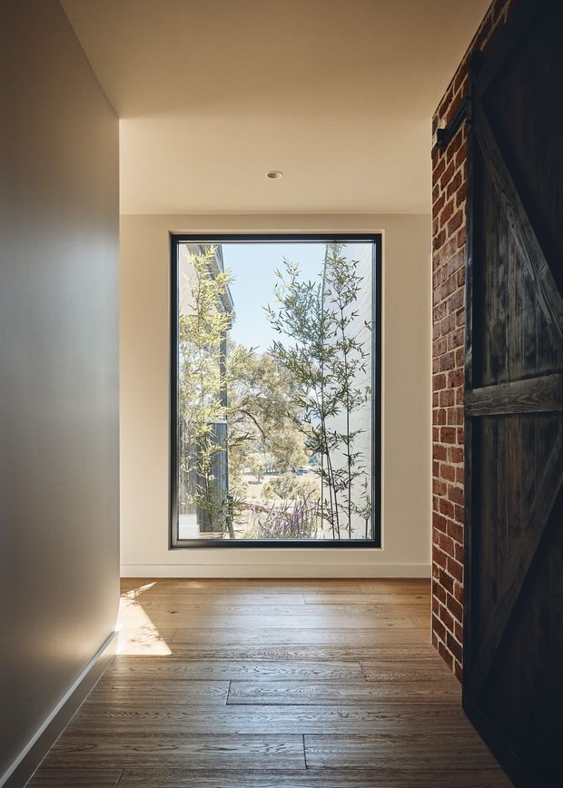 A narrow, sunlit interior passage or entry area with warm timber flooring, smooth light-painted walls and ceiling, a large black-framed window, and an exposed red-brick wall beside a dark timber barn-style door. Landscaping and a neighbouring structure are visible through the window.
