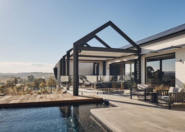 Contemporary outdoor entertaining area beside a swimming pool, with a broad timber deck, paved terrace, glass-enclosed lounge or dining space, black steel framing and a pitched gable-like roof structure. The adjacent white brick building has large glazed openings and rooftop solar panels, while a dry rural landscape extends beyond the property.