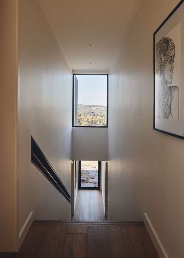 A narrow, tall interior stairway descending through a contemporary home. The stair treads and lower landing have dark timber flooring, while the walls and ceiling are painted warm off-white. Slim black-framed windows at the upper and lower levels bring in daylight and frame a distant landscape. A large monochrome framed artwork hangs on the right wall, and a dark built-in wall feature or stair railing runs along the left side.