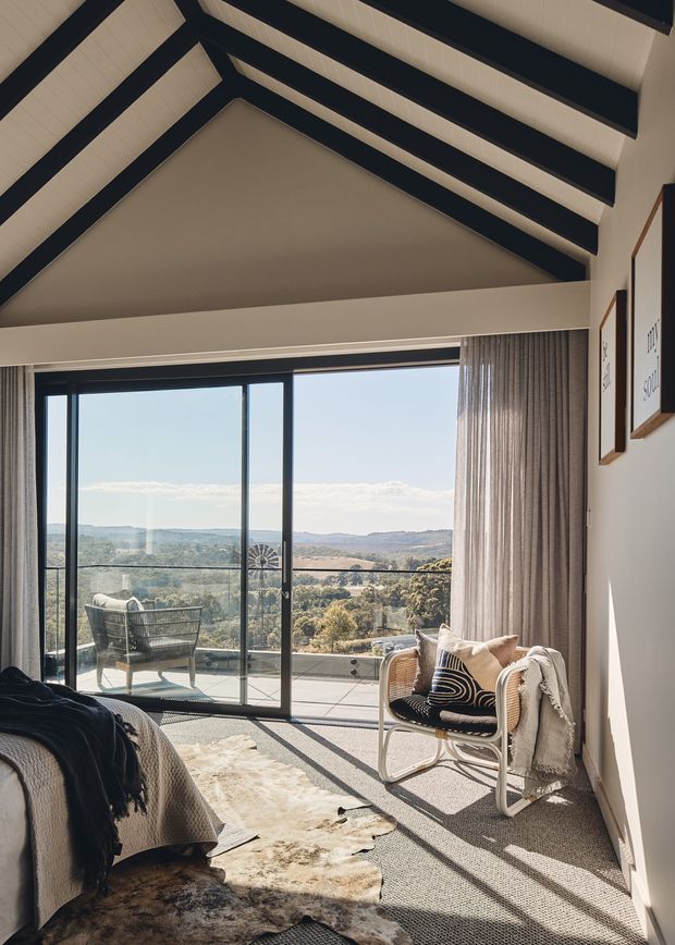 A bright bedroom with a high vaulted ceiling featuring exposed dark beams and pale ceiling panels. Full-height sliding glass doors open to a balcony or terrace with expansive rural views. The room includes a bed, woven lounge chair, textured rug, sheer curtains, and framed wall artwork. The interior has a warm, relaxed contemporary character with strong indoor-outdoor connection.