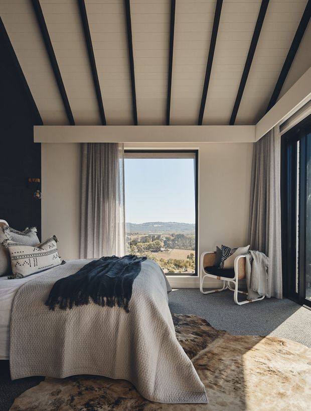A calm, contemporary bedroom with a large bed facing a picture window and expansive corner glazing. The room features a pale upholstered or textured bed covering, layered neutral bedding, dark navy-black accent walls and ceiling rafters, full-height grey curtains, woven carpet, and two light-framed lounge chairs. A rural landscape is visible through the windows.
