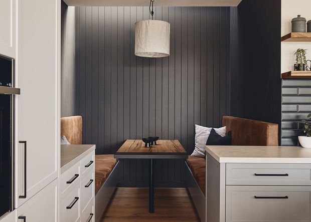 A compact built-in dining nook viewed within a contemporary kitchen. The nook features dark vertical wall panelling, a timber table with black pedestal base, upholstered bench seating and cushions, and a large cylindrical pendant light. White shaker-style cabinetry and drawers frame the space, with open shelving and additional cabinetry visible at right. The palette is predominantly white, charcoal, brown timber, and tan upholstery.