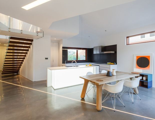 Bright open-plan contemporary kitchen and dining area with a white island, dark kitchen cabinetry, stainless-steel rangehood, timber dining table, white molded chairs, polished concrete flooring, and a striking open timber-and-glass staircase. A narrow horizontal window and clerestory-style glazing bring in natural light, while recessed and linear lighting create a clean modern interior.