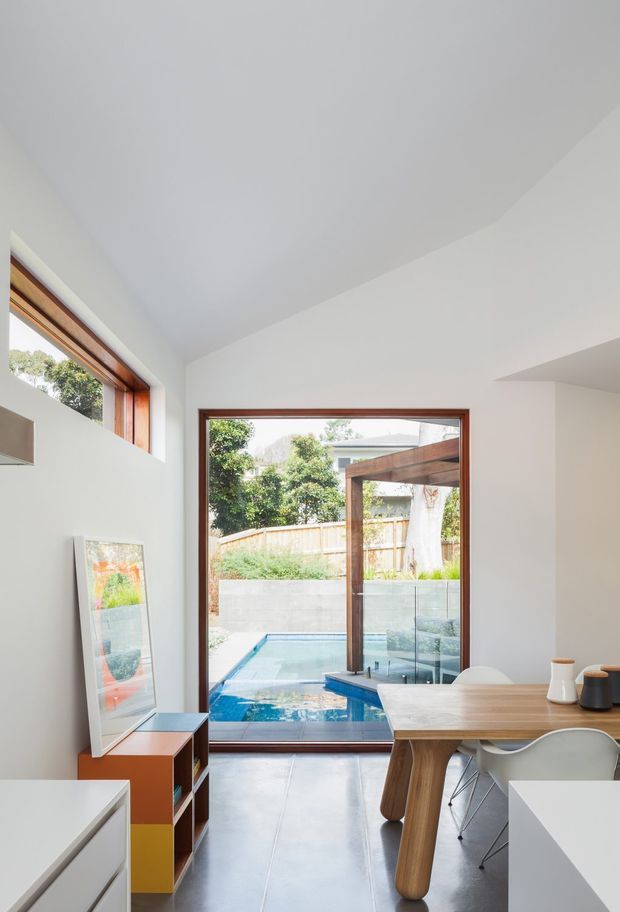 Bright contemporary dining and living area viewed from inside, with white walls and ceiling, warm timber-framed glazing, a large timber dining table with modern chairs, and a compact outdoor swimming pool visible beyond the glass doors. The room has a clean open connection to the landscaped courtyard and strong natural light.