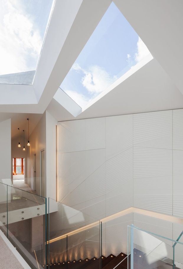 Bright contemporary interior view of a multi-level stairwell and landing, featuring expansive geometric skylights, white textured wall panelling, glass balustrades, dark timber stairs, and subtle integrated lighting. A corridor with pendant lights and a timber-framed window is visible in the background.