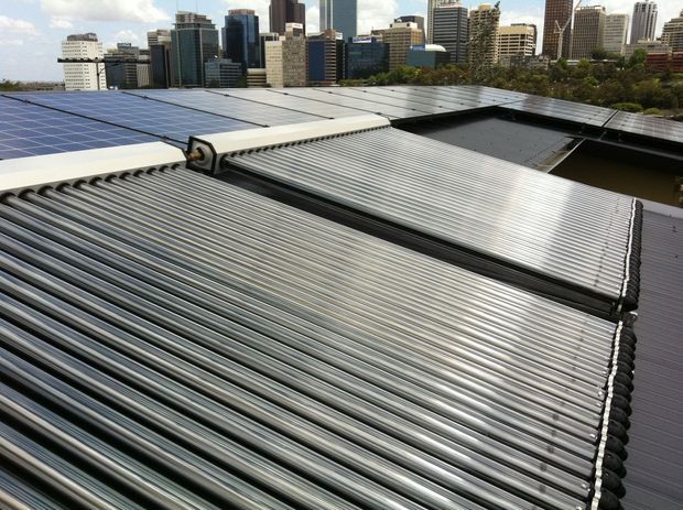 Close-up exterior view of a large corrugated metal roof fitted with extensive solar panels and a prominent bank of cylindrical solar thermal collector tubes. A dense modern city skyline and tree-lined urban setting are visible in the background.