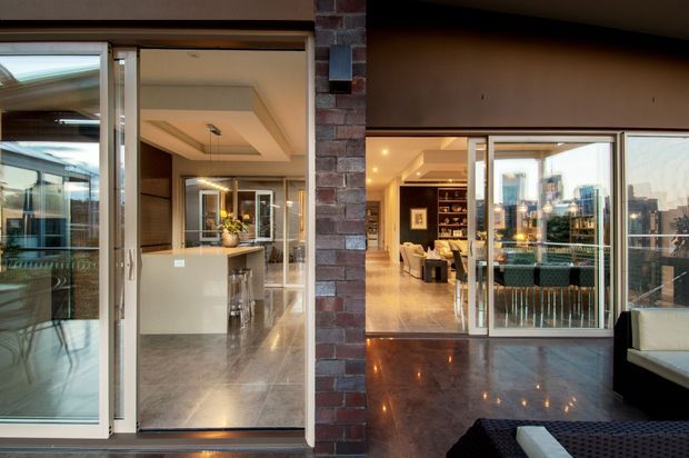 View from a covered outdoor terrace or balcony through wide glazed sliding doors into a polished, contemporary residential interior. The image shows a strong brick structural pier dividing two openings, reflective dark flooring on the terrace, full-height glass doors, and an interior with a kitchen island, lounge area, built-in shelving, and warm recessed lighting. A city skyline is visible through the glazing and reflections.