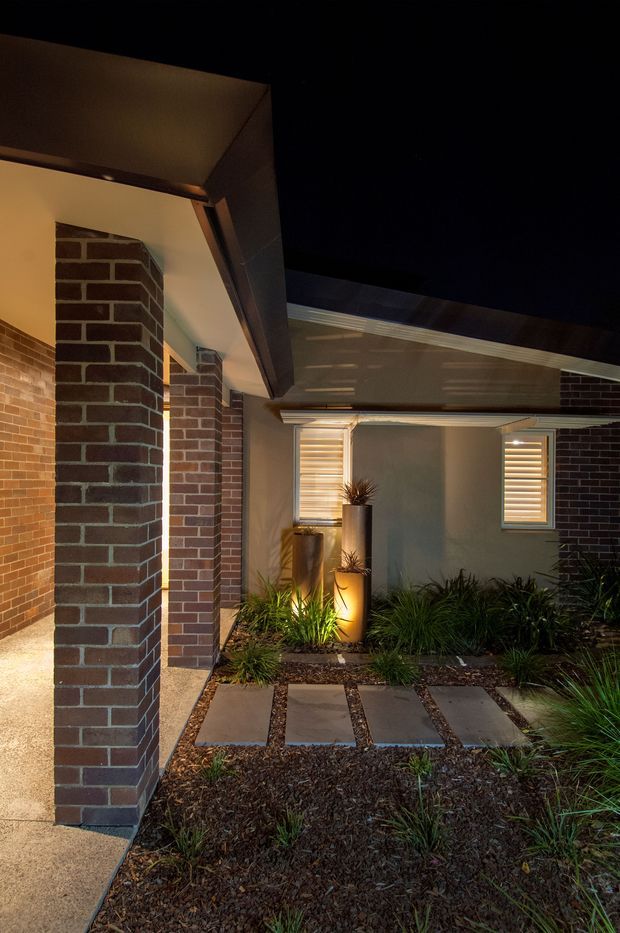 Night-time exterior view of a residential entry courtyard. Dark red-brown brick columns and walls frame a covered walkway and recessed entrance, with grey rendered wall sections, white louvred windows, and a pitched tiled roof. Rectangular concrete stepping slabs cross a landscaped garden bed filled with ornamental grasses, while warm integrated lighting illuminates the planters and facade.