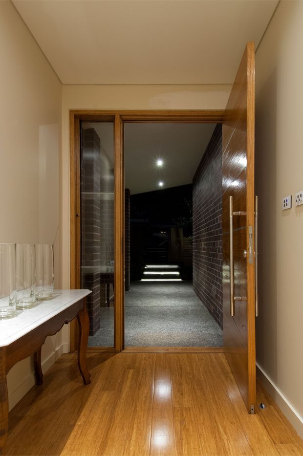 Interior view of a warm-toned entryway with polished timber flooring, cream walls and ceiling, and a timber-framed glazed doorway opening to a covered exterior walkway. A large timber door with a long vertical metal pull handle is open, while a small console table with glassware sits along the left wall. The exterior passage beyond has dark brick walls, recessed lighting, and illuminated steps.