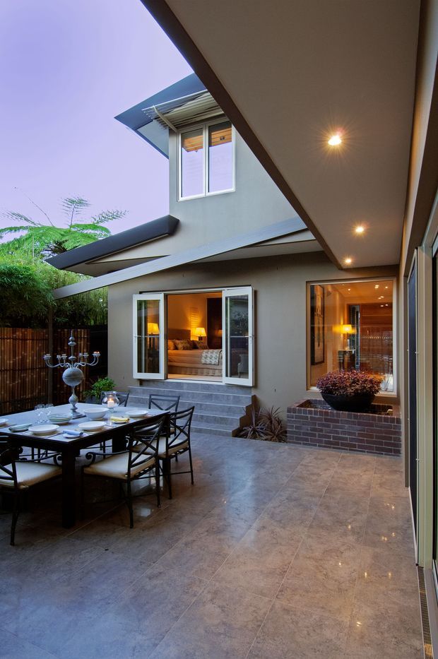 A covered courtyard or outdoor dining terrace viewed at dusk, with a large tiled floor, dining table and chairs, built-in brick planter, lush screening plants, and dramatic contemporary roof overhangs. The courtyard opens toward a warmly lit living room through folding or bi-fold windows and doors, while recessed downlights illuminate the covered area.