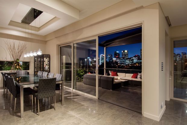 A refined open-plan interior at night, with a formal dining area in the foreground and a lounge opening onto a covered terrace. Large sliding glass doors frame illuminated city high-rises, while polished stone-look flooring, neutral walls, a glass dining table, upholstered chairs, and a cluster pendant light create an elegant urban setting. A mirrored ceiling recess adds visual height and reflects the dining area.