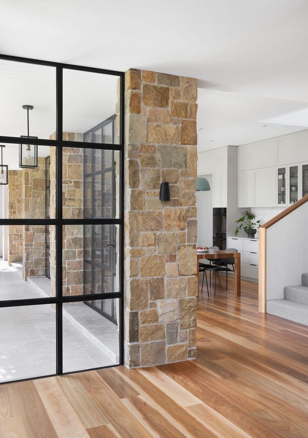 Bright contemporary residential interior showing an open-plan dining and kitchen area beside a stairway. A prominent exposed natural stone structural wall is paired with black steel-framed glazed partitions, white built-in cabinetry, timber flooring, and a carpeted staircase with timber handrail. The glazing opens visually toward a light-filled courtyard or adjacent space.