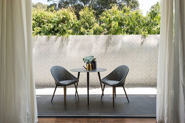 A bright, refined indoor-outdoor seating area viewed through open floor-to-ceiling curtains. Two sculptural dark chairs face each other across a small round table with a potted plant and decorative objects. The terrace has large dark tiled flooring and a pale geometric patterned privacy wall, with leafy trees visible beyond.