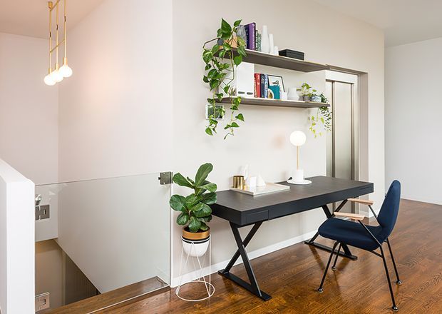 Bright contemporary home office positioned beside an open stairwell. The room features a dark built-in or wall-mounted shelving unit with books and plants, a large dark desk with angled metal legs, a navy upholstered chair, a small table lamp, pendant lighting, white walls, and warm timber flooring. A frameless glass stair balustrade and doorway are visible in the background.
