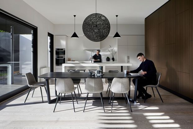 A bright, open-plan contemporary kitchen and dining space with a long dark dining table surrounded by upholstered chairs. A white kitchen with island and integrated appliances sits behind the dining area, while a full-height dark timber feature wall and expansive black-framed glazing define the room. Two black pendant lights and a large spherical sculptural pendant hang above the table. Natural daylight creates strong linear shadows across the pale timber floor.