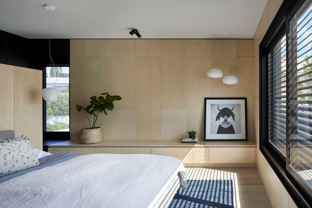 A bright, minimalist bedroom with a low platform bed in the foreground, pale timber wall panelling and integrated built-in storage across the back wall. Large black-framed windows with horizontal blinds fill the right side, casting strong linear shadows across the timber floor. The room includes restrained pendant and wall lighting, a potted plant, and framed artwork.