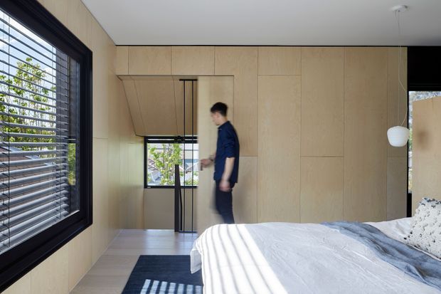 Bright contemporary bedroom with full-height light timber wall panelling and integrated cabinetry, a black-framed internal glass door, large black-framed louvred window, and a neatly made bed in white and pale grey bedding. The room has a restrained, clean-lined palette with strong daylight and views to greenery through the glazing.