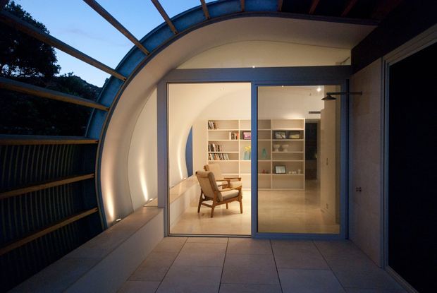 An evening view from a covered outdoor terrace or balcony into a softly lit contemporary interior. The terrace has large square floor tiles, a curved translucent roof and structural framing, and dark screened side panels. Through full-height sliding glass doors, a calm study or living area is visible with built-in white shelving, books, display objects, a timber lounge chair, and polished light flooring.