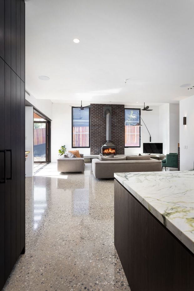 Bright open-plan contemporary interior viewed from the kitchen toward a living room. The space features a large dark timber kitchen island with a pale veined stone benchtop, polished terrazzo flooring, white walls and ceiling, a dark exposed brick fireplace wall with a suspended wood-burning fire, neutral sofas, ceiling fans, recessed lighting, and large glazed openings to an outdoor area.