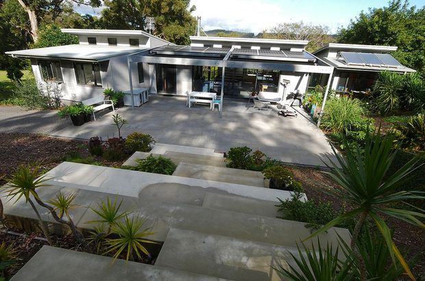 A low-profile, single-storey modern residence viewed from a landscaped garden. The home features white rendered walls, extensive dark-framed glazing, multiple shallow roof planes with skylights, and a broad covered outdoor terrace furnished with outdoor seating. Concrete steps and paving lead through drought-tolerant, tropical-style planting toward the house. A wooded landscape and distant hills are visible beyond.