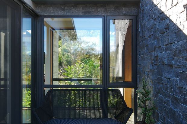 A compact outdoor or semi-enclosed seating nook framed by dark metal window and door glazing, with a black woven lounge chair in the foreground. The surrounding walls are clad in rough dark stacked stone, while abundant greenery is visible through the glazing. Strong sunlight creates high contrast across the textured masonry and frames.