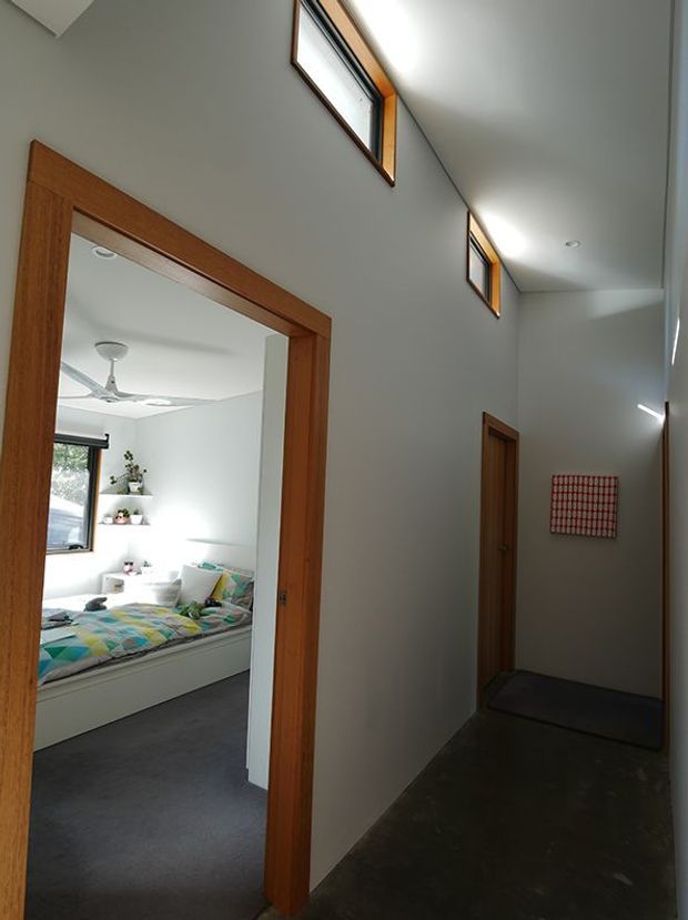 A narrow interior hallway with dark flooring, white walls and timber door frames. High-level clerestory windows bring daylight into the corridor, while an open doorway reveals a bright bedroom with a built-in bed, colourful bedding, shelving and a ceiling fan. A closed timber door and small wall artwork line the hall.