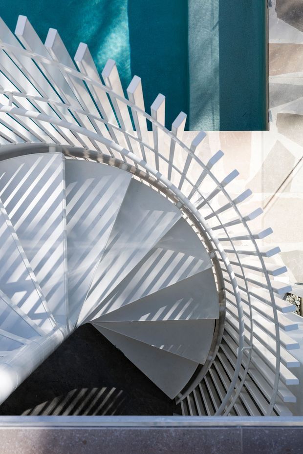 Top-down architectural view of a white spiral stairway with curved metal railings and closely spaced vertical balusters, casting strong linear shadows across the steps. A vivid turquoise surface and pale geometric paving are visible adjacent to the stairwell, creating a high-contrast graphic composition.